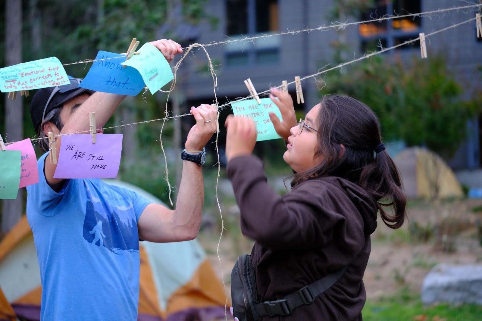 Two people stand at and actively engage with what appears to be a clothesline. Colourful pieces of paper
    with text written on them are pinned to the clothesline.  On one piece of paper can be seen the hand-written
    text "WE ARE STILL HERE" while on others can be seen the words "Know that..." and "You are your ancestors".