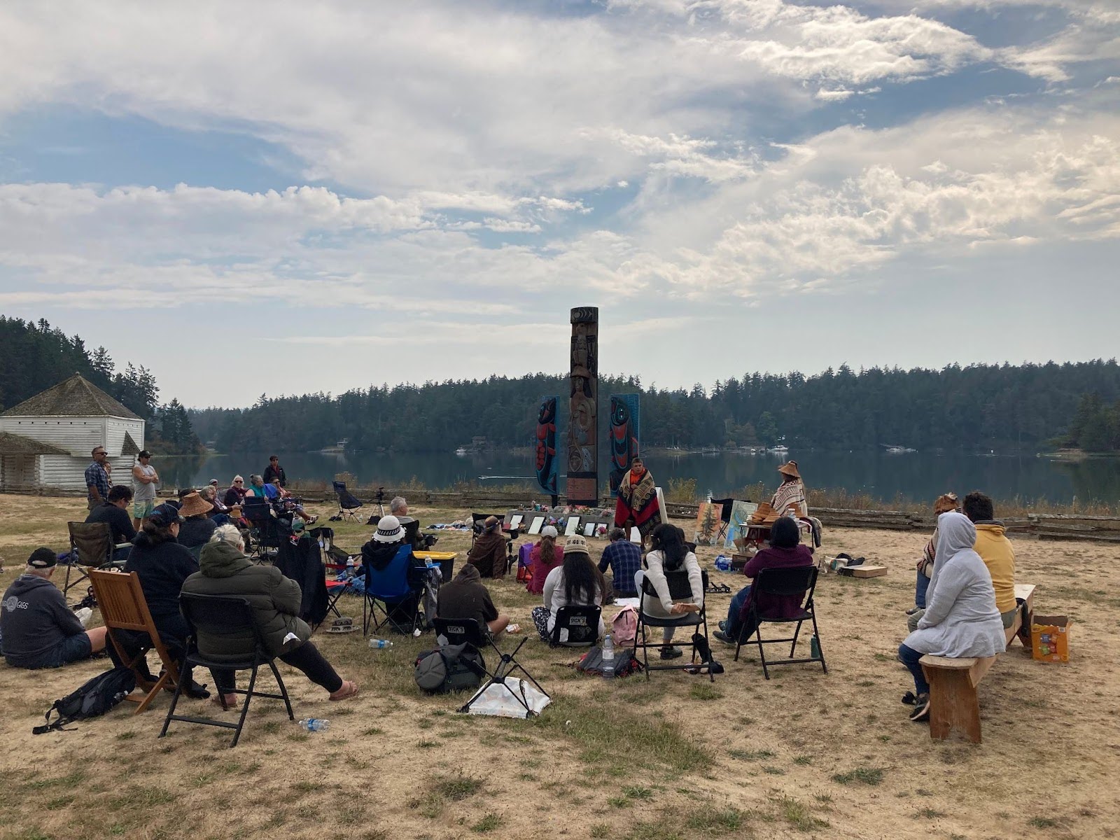 A group of approximately 25 people stand and sit in a grassy area by the water in front of 3 carved and
    painted Strait Salish story poles. A man wearing a blanket over his shoulders stands in front of the group and
    appears to be talking, while a woman in a woven Salish blanket and cedar hat stands near him listening.
    A white clapboard building can be seen in the background.