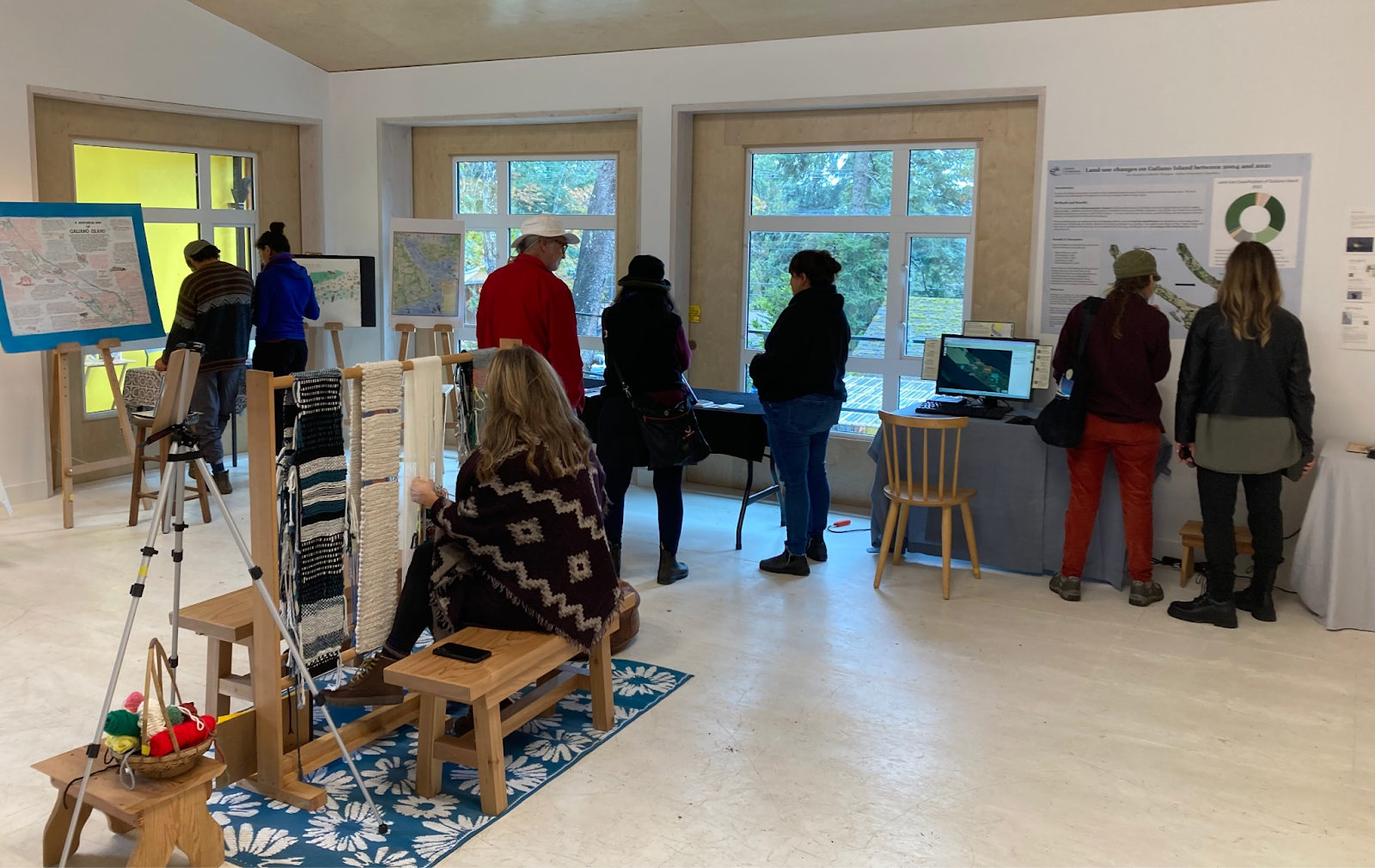 A number of people stand around in the gallery space, looking at the various parts of the exhibit and talking
    together. One person sits at the loom, weaving.