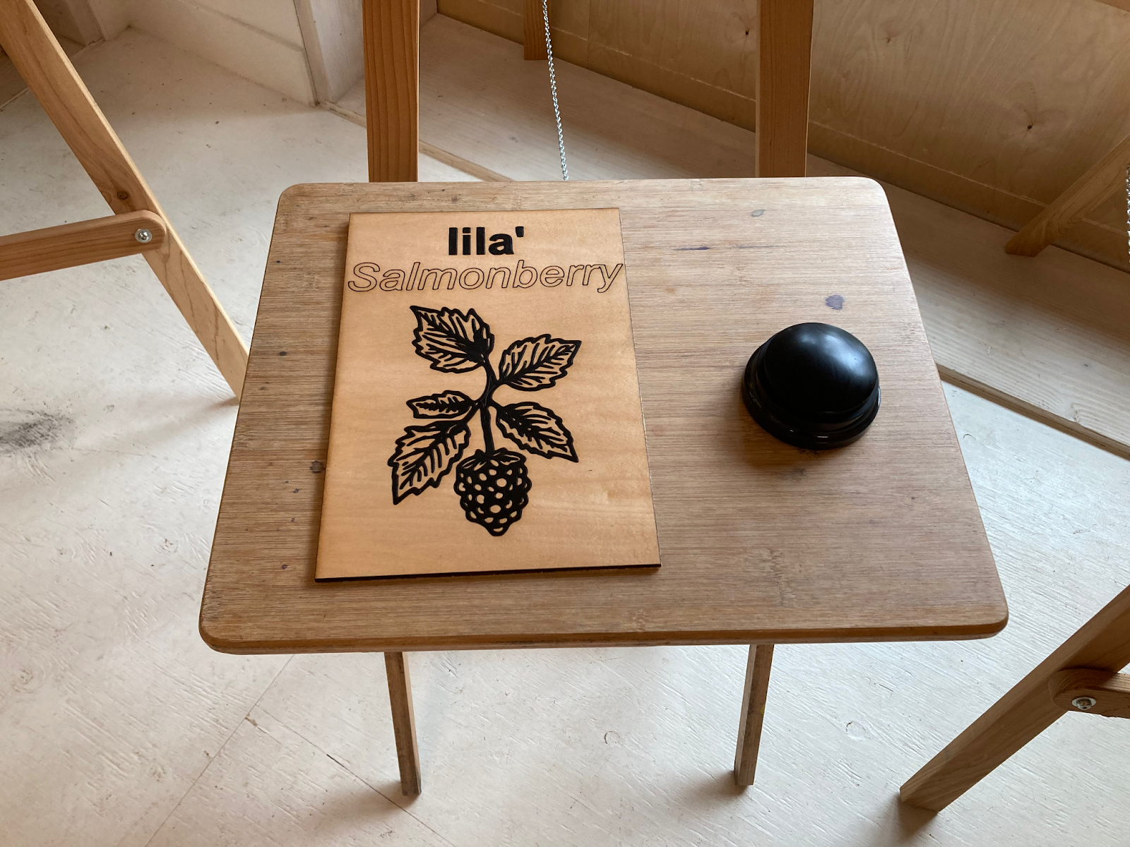 A small wooden table with a wooden panel lying on it, with the word lila' in bold at the top, Salmonberry
    underneath, and an image of a salmonberry in the centre. Beside the panel is a large, round, black button.